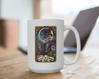 May include: White ceramic mug featuring a tarot card design with a skeleton in a purple suit and top hat. The card has a gold border and the text "F*ck it, I'm already dead". The mug is on a wooden surface.