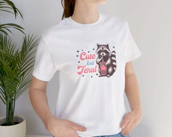 Op de afbeelding: Wit T-shirt met een illustratie van een wasbeer die een roze drankje drinkt, en de tekst "Cute but Feral". De wasbeer is zwart en grijs, het drankje is roze. De tekst is roze en wit. Een casual kledingstuk.