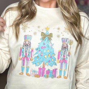 May include: A cream colored long-sleeved sweatshirt with a festive design featuring three nutcracker figures standing in front of a blue and green Christmas tree with gold accents and presents. The design is printed on the front of the sweatshirt.