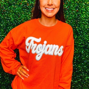 Puede incluir: Una sudadera naranja brillante con la palabra "Trojans" en letras blancas en la parte delantera.