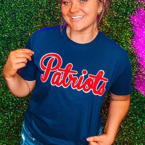Op de afbeelding: Een vrouw draagt een donkerblauw T-shirt met het woord "Patriots" in rode letters. Ze staat voor een groene muur met een roze neonbord waarop "South & Main" staat.