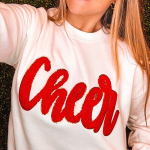 May include: White sweatshirt with a red and gold glitter "Cheer" graphic.