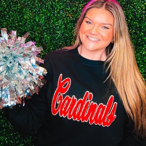 May include: A woman wearing a black sweatshirt with the word "Cardinals" in red lettering. She is holding a silver pom-pom in her left hand. The background is a green wall with a pink neon sign that says "South & Main."