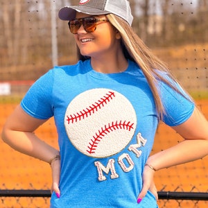 May include: A light blue t-shirt featuring a large baseball design with red stitching and the word "MOM" in red letters. The person is wearing a baseball cap with a baseball logo and sunglasses.