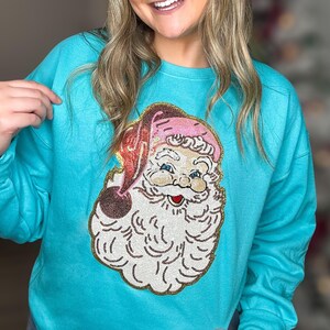 Może przedstawiać: Turkusowa bluza z haftowanym wzorem Świętego Mikołaja. Święty Mikołaj ma różowy kapelusz i białą brodę.