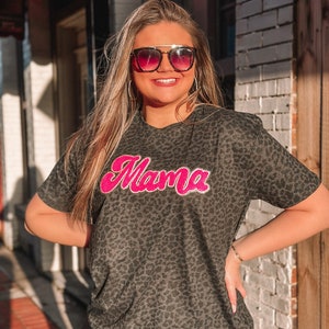 May include: A dark grey leopard print t-shirt with the word "Mama" in bright pink, outlined in white. The shirt has short sleeves and a crew neck. The person in the image is wearing sunglasses and hoop earrings.
