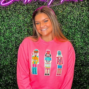 Op de afbeelding: Roze trui met drie geborduurde notenkrakerfiguren. De notenkrakers dragen kleurrijke kostuums en hoeden. De trui heeft een ronde hals en lange mouwen.