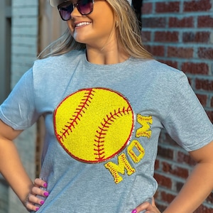 May include: A grey t-shirt featuring a yellow and red softball graphic and the word "MOM" in glitter. The model is wearing a black and grey baseball cap with a white mesh back.
