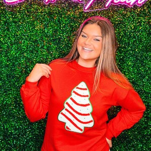 May include: A red sweatshirt with a white and red striped Christmas tree design. The sweatshirt is worn by a person standing in front of a green wall with a pink neon sign that says "South & Main".