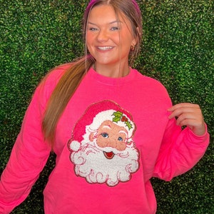 Może przedstawiać: Jasnoróżowa bluza z nadrukiem Świętego Mikołaja z przodu. Wzór Świętego Mikołaja obejmuje białą brodę, różowe policzki i czerwony kapelusz z ostrokrzewem. Neonowy znak powyżej głosi "South & Main".