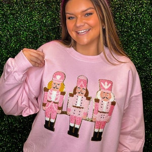 Könnte beinhalten: Rosafarbener Pullover mit einem festlichen Design aus drei rosafarbenen Nussknackern mit goldenen Akzenten. Der Pullover hat einen Rundhalsausschnitt und lange Ärmel.