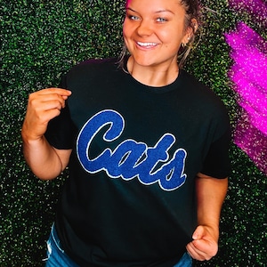 May include: A black t-shirt with a blue "Cats" graphic. The shirt is worn by a person standing in front of a green wall with a pink neon sign that says "South & Main".