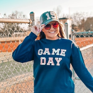 Può includere: Una felpa blu navy con la scritta "GAME DAY" in lettere bianche bordate da cuciture rosse. Le lettere sono fatte per assomigliare a palle da baseball. La persona indossa un cappello da baseball verde e nero con il numero "19" sopra.