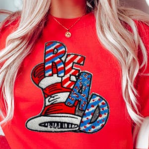 May include: A red sweatshirt with the word "READ" in a whimsical font, featuring a hat design with red and white stripes and blue polka dots. The sweatshirt is a vibrant red color.
