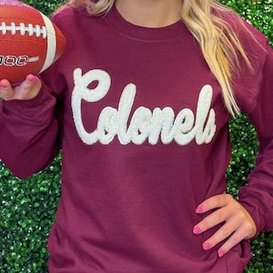 May include: A maroon long-sleeved sweatshirt with a white, fuzzy, cursive "Colonels" graphic. A person is holding a red and white football.