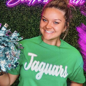 May include: A woman wearing a green t-shirt with the word "Jaguars" in white glitter lettering. She is holding a silver pom-pom.
