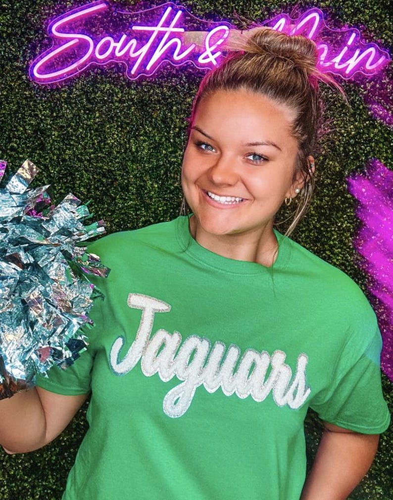 May include: A woman wearing a green t-shirt with the word "Jaguars" in white glitter lettering. She is holding a silver pom-pom.