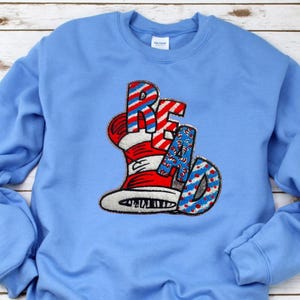May include: Light blue sweatshirt with the word "READ" in a whimsical design. The letters are red and white striped, with a hat design. The "A" and "D" are blue with white polka dots. The sweatshirt is a solid light blue color.