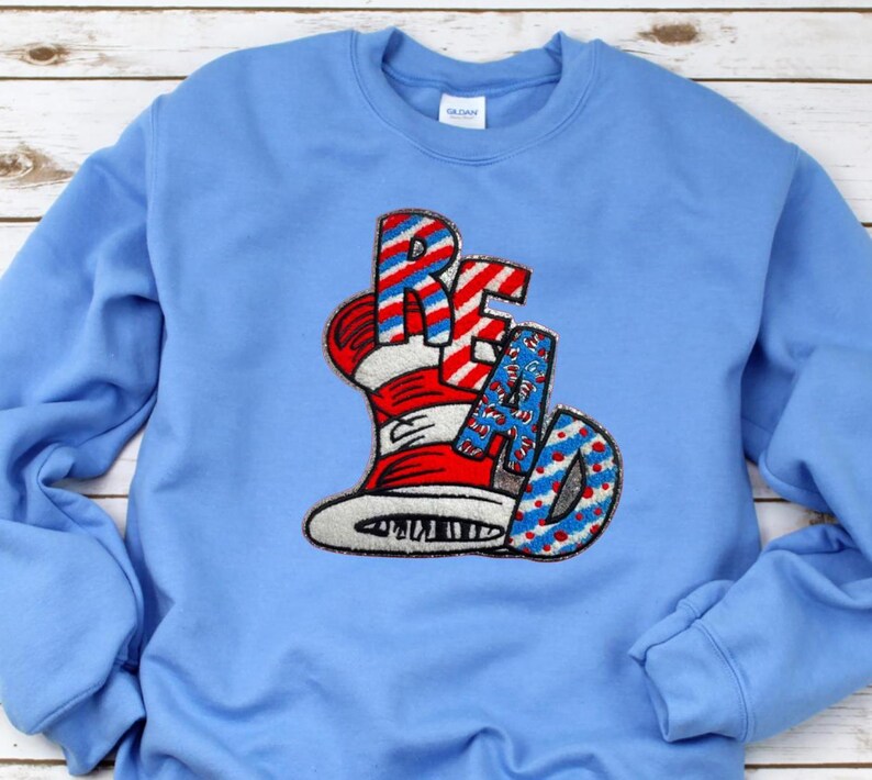 May include: Light blue sweatshirt with the word "READ" in a whimsical design. The letters are red and white striped, with a hat design. The "A" and "D" are blue with white polka dots. The sweatshirt is a solid light blue color.