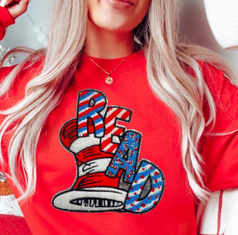 May include: A red sweatshirt with the word "READ" in a whimsical font, featuring a hat design with red and white stripes and blue polka dots. The sweatshirt is a vibrant red color.