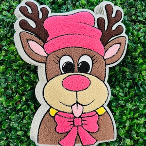 Puede incluir: Una cara de reno de peluche con un gorro y un lazo rosas. El reno tiene cuernos marrones, un hocico beige y una nariz rosa. El contorno es de color gris claro y el fondo es verde.