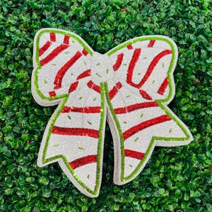 Puede incluir: Un gran lazo decorativo con una base de lentejuelas blancas, acentos de rayas rojas y un borde de lentejuelas verdes. El lazo es probablemente para la decoración navideña o de regalos.