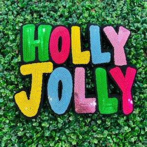 Puede incluir: Un parche de lentejuelas de colores con las palabras "HOLLY JOLLY" en letras grandes. Las letras son de varios colores, incluyendo verde, rosa, amarillo y azul, sobre un fondo negro. El parche está sobre un fondo de follaje verde.