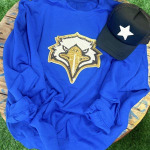 Könnte beinhalten: Ein königsblauer Pullover mit einem goldenen Pailletten-Adlerkopf-Design. Der Pullover hat lange Ärmel und einen Rundhalsausschnitt.