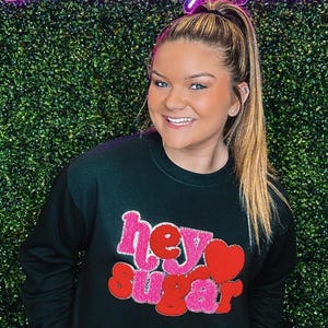 K&ouml;nnte beinhalten: Ein schwarzer Pullover mit einem pinkfarbenen und roten "hey sugar"-Motiv auf der Vorderseite. Das Motiv ist aus flauschigem Material und hat die Form eines Herzens.