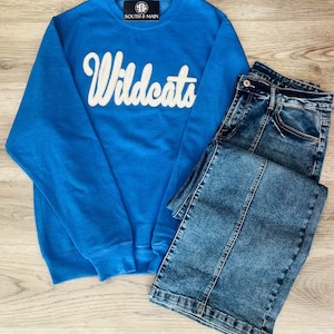 May include: A blue sweatshirt with the word "Wildcats" in white script on the front. The sweatshirt is paired with a pair of blue denim jeans.