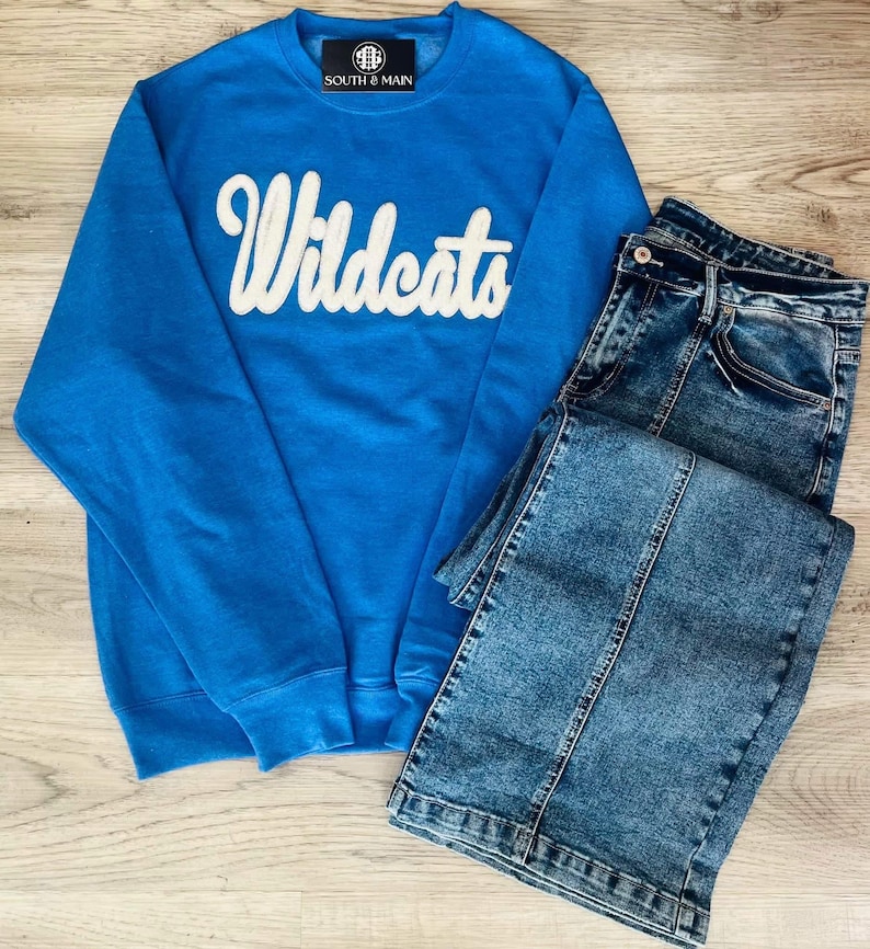 May include: A blue sweatshirt with the word "Wildcats" in white script on the front. The sweatshirt is paired with a pair of blue denim jeans.