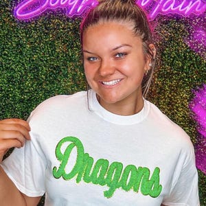 Peut inclure: Un t-shirt blanc avec un graphisme vert "Dragons". Le t-shirt est port&eacute; par une personne aux cheveux bruns et un chignon. L'arri&egrave;re-plan est un mur vert avec un panneau n&eacute;on rose qui dit "South Main".