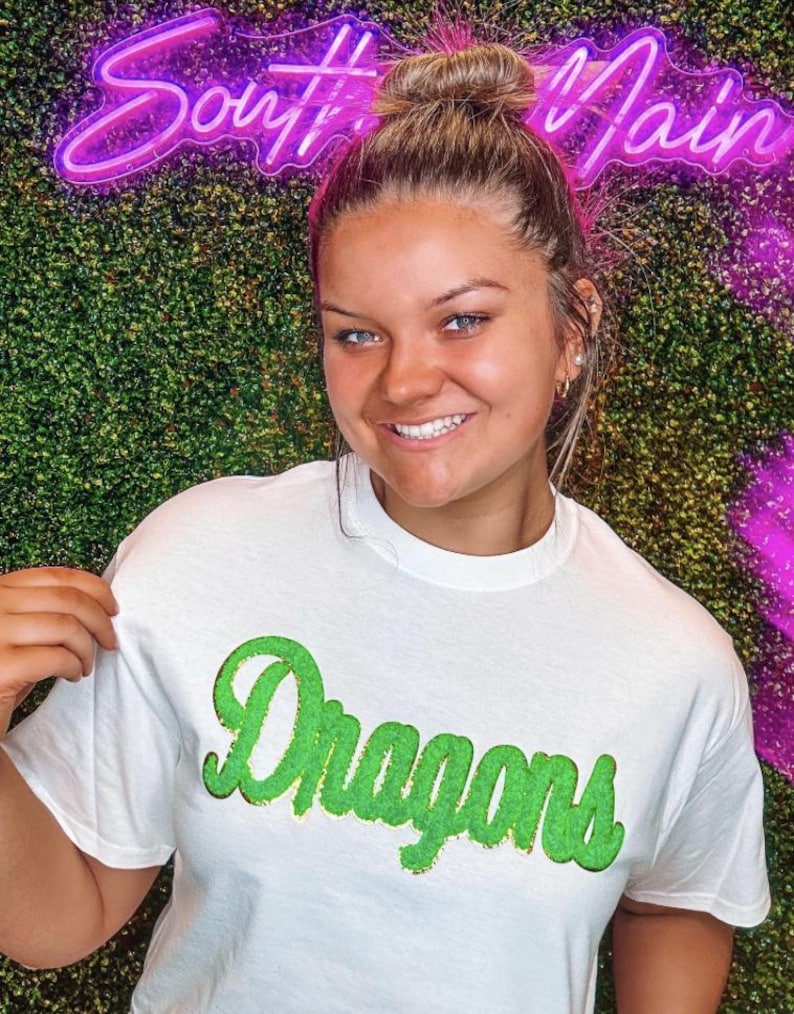 Peut inclure: Un t-shirt blanc avec un graphisme vert "Dragons". Le t-shirt est port&eacute; par une personne aux cheveux bruns et un chignon. L'arri&egrave;re-plan est un mur vert avec un panneau n&eacute;on rose qui dit "South Main".