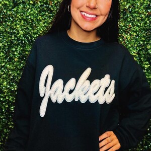 Puede incluir: Sudadera negra con un gr&aacute;fico blanco y brillante de "Jackets".