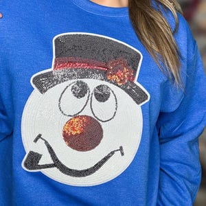 Può includere: Una felpa blu reale con un viso di pupazzo di neve bianco con occhi neri, un naso rosso e un cappello a cilindro nero con una fascia rossa e un fiore rosso.