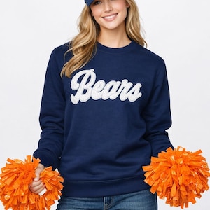 May include: A navy blue sweatshirt with the word "Bears" in white script. The person is wearing a navy blue baseball cap with a white paw print and holding two orange pom-poms. The person is also wearing blue jeans.