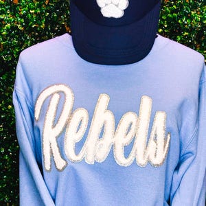 Puede incluir: Una sudadera azul claro con la palabra "Rebels" en blanco con un contorno plateado. La sudadera tiene mangas largas y cuello redondo. Una gorra de camionero de malla negra con una huella de pata blanca está encima de la sudadera.