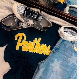 May include: A pair of grey trainers with silver glitter detailing and white star motifs. The trainers are displayed with a navy blue t-shirt featuring the word "Panthers" in yellow script, and light blue denim jeans with white star patterns.
