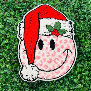 May include: A decorative rug featuring a smiley face wearing a Santa hat. The face has black eyes and a pink leopard print pattern. The Santa hat is red with a white trim and a sprig of holly with red berries.