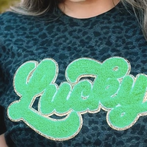 May include: A black and green leopard print t-shirt with a green and gold glitter "Lucky" graphic.