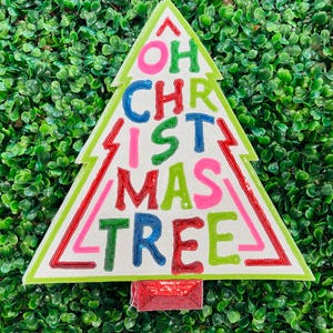 May include: A Christmas tree-shaped decoration with the words "OH CHRISTMAS TREE" in colorful, beaded letters. The tree has a white background, green outline, and red accents. The base is a red rectangle.