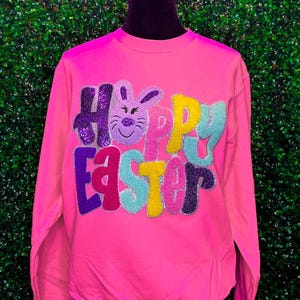 Könnte beinhalten: Rosafarbener Langarm-Pullover mit einem glitzernden "Frohe Ostern"-Design mit einem Hasen.