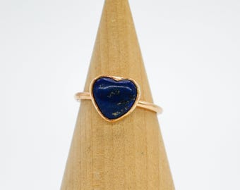 May include: A heart-shaped ring with a blue lapis lazuli stone set in a gold-toned band. The ring is displayed on a wooden ring holder.