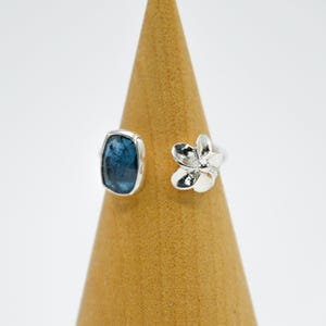 Può includere: Un anello d'argento con una pietra preziosa blu e un design a fiore. L'anello è su un espositore in legno.