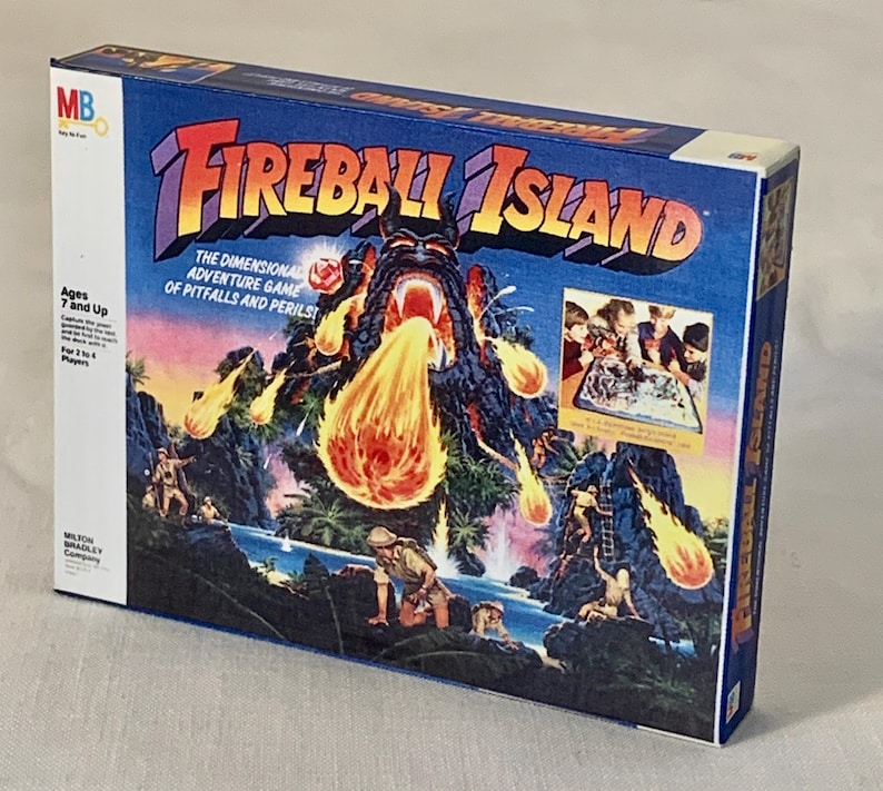 Może przedstawiać: Pudełko gry planszowej Fireball Island. Pudełko zawiera żywą ilustrację wulkanicznej wyspy z wybuchem ognia. Tekst na pudełku zawiera "Fireball Island" i "The Dimensional Adventure Game of Pitfalls and Perils." Wiek 7 lat i więcej.