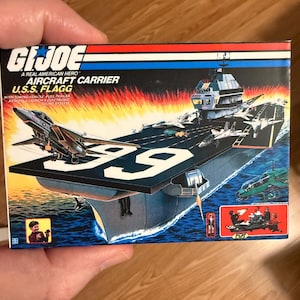May include: A vintage G.I. Joe U.S.S. Flagg aircraft carrier toy box. The box features an illustration of the carrier with fighter jets, helicopters, and the number 99 on the deck. The box has the text "A REAL AMERICAN HERO".