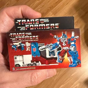 May include: A vintage Transformers Ultra Magnus toy box. The box features a red, white, and blue robot illustration and the text "TRANSFORMERS". The box also shows a white truck transforming into a robot. The text "MORE THAN MEETS THE EYE!" is also visible.
