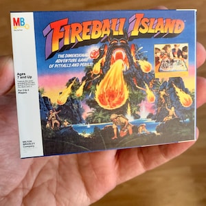 Może przedstawiać: Vintage pudełko gry planszowej Fireball Island. Pudełko zawiera kolorową ilustrację wulkanicznej wyspy z erupcją ognia. Tekst zawiera "Fireball Island" i "The Dimensional Adventure Game of Pitfalls and Perils!". Dla os&oacute;b w wieku 7 lat i starszych.