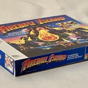 Może przedstawiać: Vintage pudełko gry planszowej Fireball Island. Pudełko jest gł&oacute;wnie niebieskie z tytułem gry w kolorze pomarańczowym i ż&oacute;łtym. Grafika na pudełku przedstawia wulkaniczną wyspę z postaciami i kulami ognia. Gra jest opisana jako "Wymiarowa gra przygodowa pułapek i niebezpieczeństw."