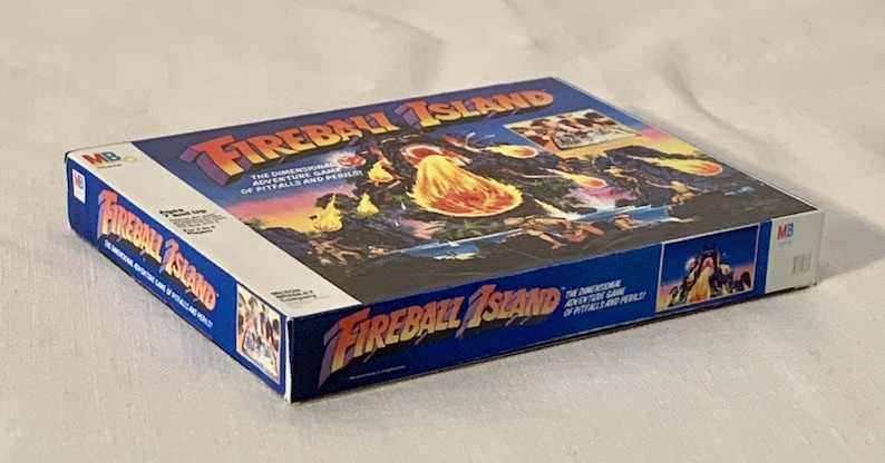 Może przedstawiać: Vintage pudełko gry planszowej Fireball Island. Pudełko jest gł&oacute;wnie niebieskie z tytułem gry w kolorze pomarańczowym i ż&oacute;łtym. Grafika na pudełku przedstawia wulkaniczną wyspę z postaciami i kulami ognia. Gra jest opisana jako "Wymiarowa gra przygodowa pułapek i niebezpieczeństw."